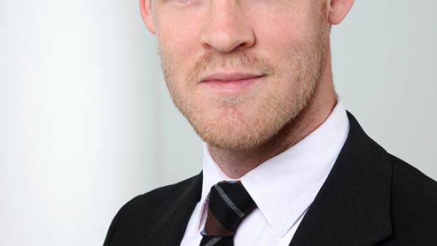 Ein Mann mit blonden Haaren und Bart trägt einen schwarzen Anzug mit Krawatte.