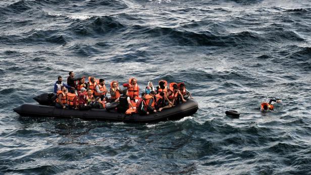 Ein überfülltes Schlauchboot mit Menschen in Schwimmwesten auf unruhiger See. Eine Person schwimmt im Wasser.