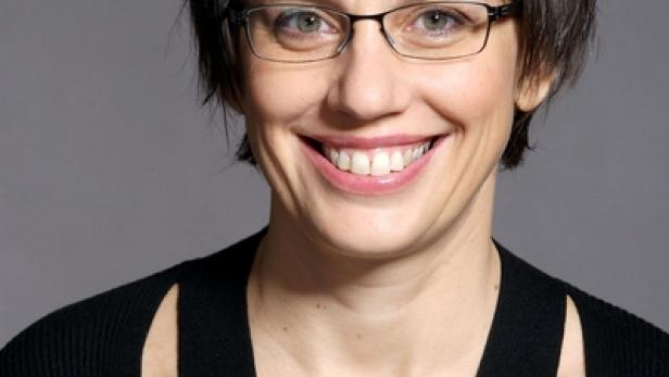 Eine lächelnde Frau mit Brille und kurzem, dunklem Haar vor grauem Hintergrund.