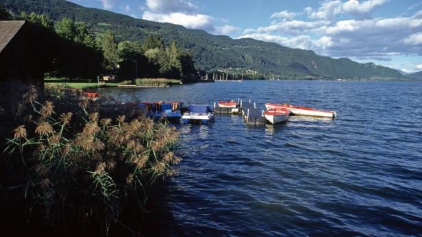 Mehrere Boote liegen an einem kleinen Steg auf einem See vor einer bewaldeten Hügellandschaft.
