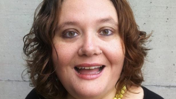 Eine Frau mit braunen, lockigen Haaren und einer gelben Halskette lächelt.