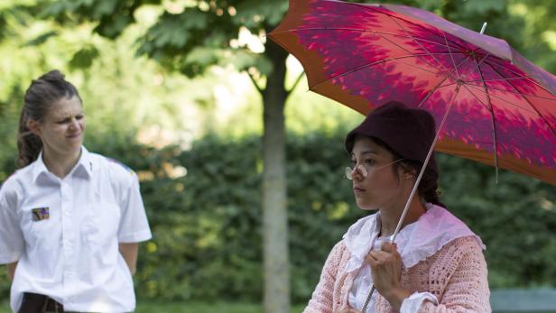 Eine junge Frau mit Hut und Schirm, im Hintergrund eine weitere Person.