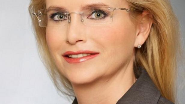 Eine blonde Frau mit Brille und grauem Blazer blickt in die Kamera.