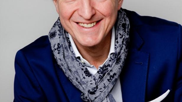 Ein lächelnder Mann mit blauem Sakko und gemustertem Schal vor hellem Hintergrund.
