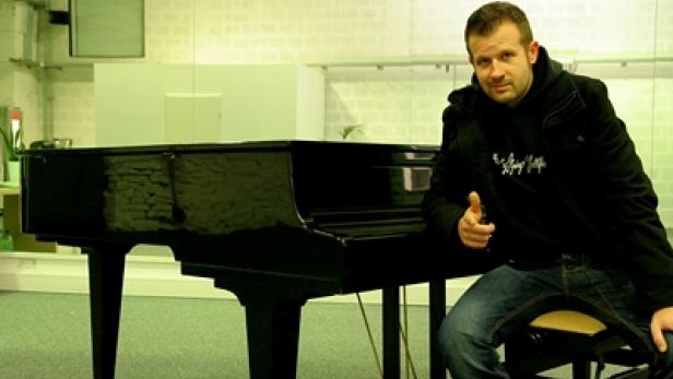 Ein Mann sitzt neben einem schwarzen Klavier und zeigt mit dem Finger auf die Kamera.