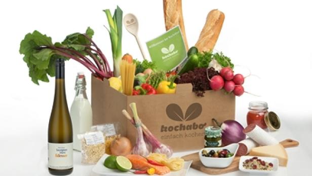 Eine Kochbox mit frischen Zutaten, darunter Gemüse, Pasta, Brot, Wein und Aufschnitt.