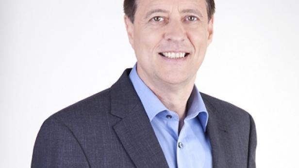 Ein lächelnder Mann in einem blauen Hemd und einem grauen Sakko vor einem weißen Hintergrund.