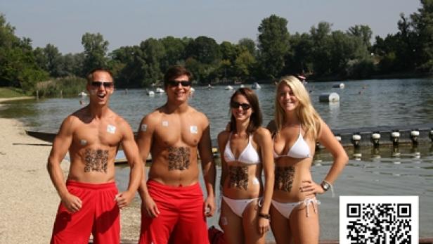 Vier Personen in Badebekleidung stehen am Ufer eines Sees und haben QR-Codes aufgemalt.
