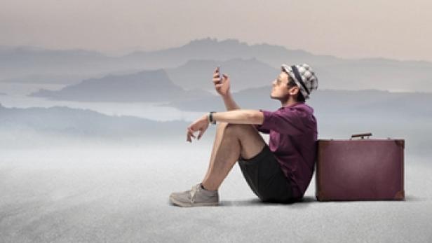 Ein Reisender mit Hut sitzt neben seinem Koffer und blickt auf sein Handy.