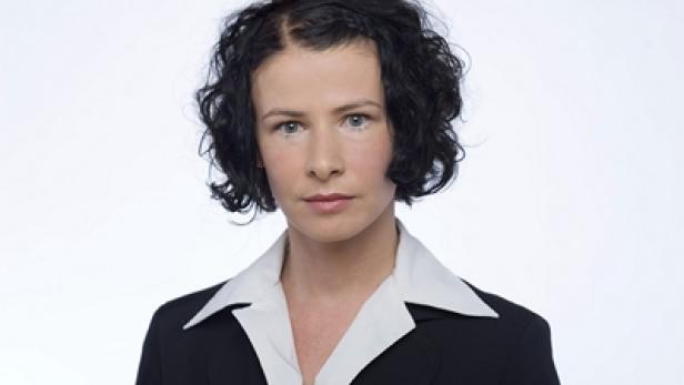 Eine Frau mit dunklen, lockigen Haaren und einem schwarzen Blazer vor einem weißen Hintergrund.