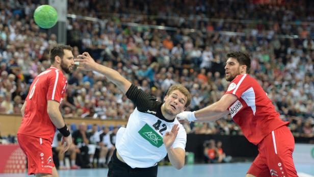 Ein Handballspieler im weißen Trikot wirft den Ball, während zwei Gegenspieler ihn beobachten.