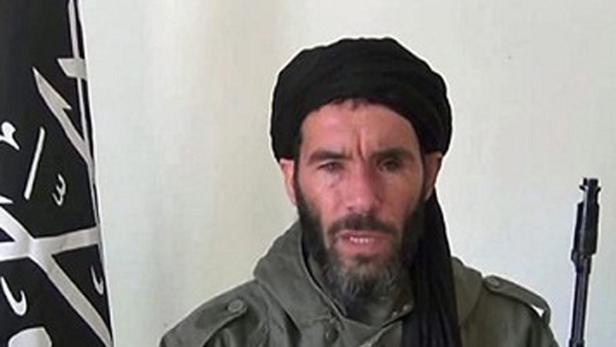 Ein Mann mit Bart und Turban steht vor einer Flagge und hält ein Gewehr.