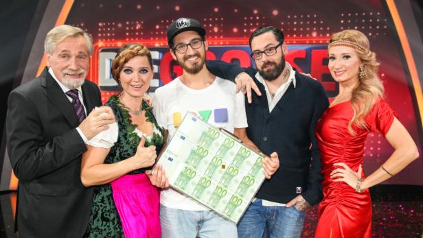 Fünf Personen posieren mit einem Koffer voller 100-Euro-Scheine auf einer Bühne.