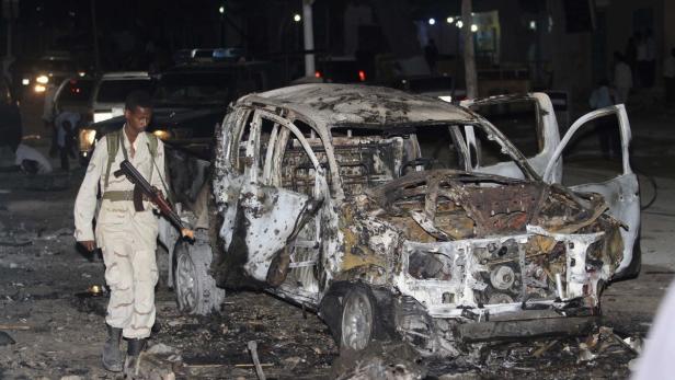 Ein Soldat mit Gewehr steht vor einem ausgebrannten Autowrack.