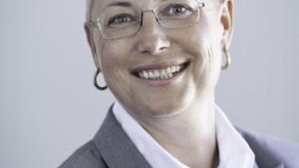 Eine lächelnde Frau mit Brille und kurzem Haar trägt ein graues Jackett.