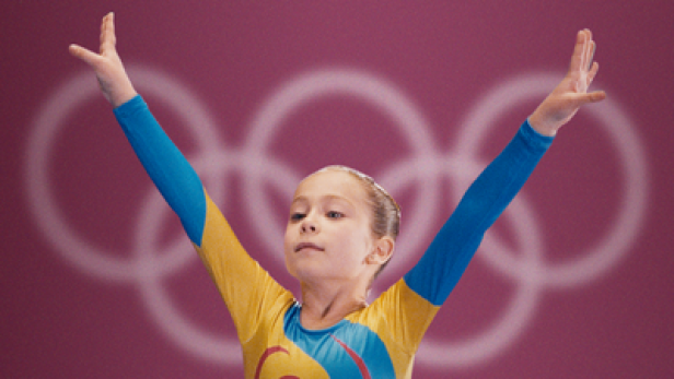 Eine junge Turnerin jubelt vor dem Hintergrund der olympischen Ringe.