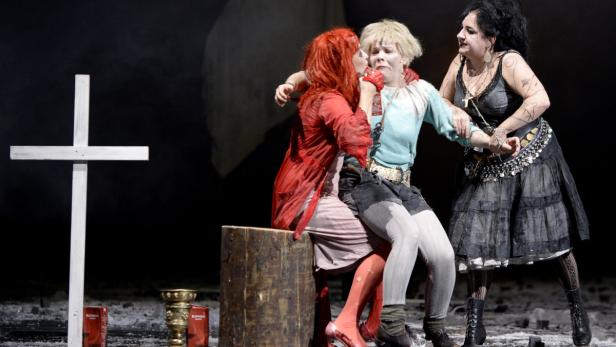 Drei Darstellerinnen in Kostümen auf einer Bühne mit einem Kreuz und Requisiten.
