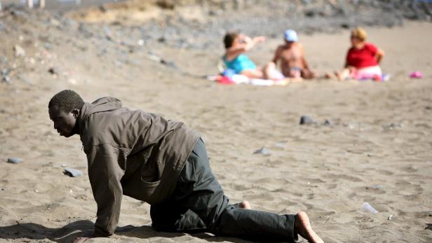 Ein Mann krabbelt barfuß am Strand, während sich im Hintergrund andere Leute sonnen.
