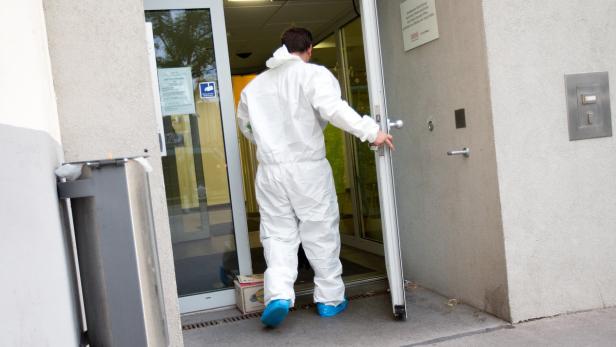 Eine Person in einem weißen Schutzanzug und blauen Überschuhen betritt ein Gebäude.