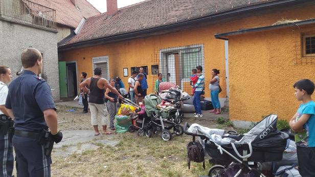 Eine Gruppe von Menschen steht vor einem gelben Gebäude mit Kinderwagen und Gepäck.