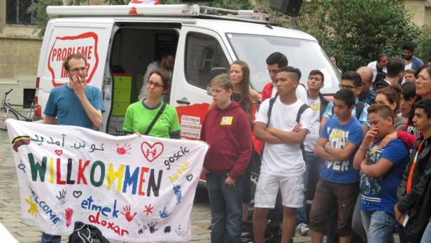 Eine Gruppe von Menschen hält ein Transparent mit der Aufschrift „Willkommen“ in verschiedenen Sprachen.