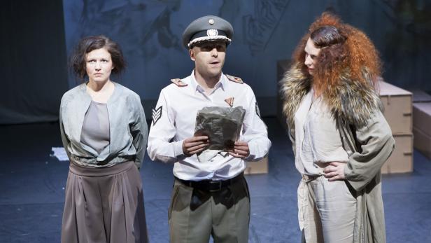 Drei Schauspieler stehen auf einer Bühne, ein Mann in Uniform liest eine Nachricht vor.