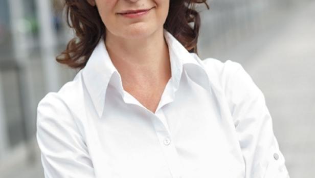 Eine Frau mit braunen Haaren und weißem Hemd steht mit verschränkten Armen da.