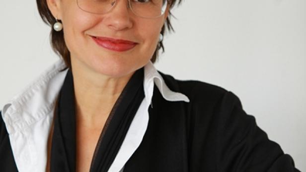 Eine Frau mit Brille, dunklem Haar und Blazer sitzt an einem Tisch.