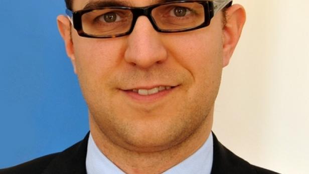 Ein Mann mit Brille, Anzug und orange-gestreifter Krawatte lächelt in die Kamera.
