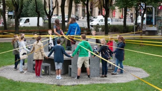 Eine Gruppe von Kindern umsteht einen Steintisch, verbunden durch gelbe Bänder.