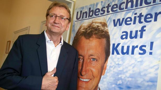 Ein Mann mit Brille posiert vor einem Wahlplakat mit der Aufschrift „Unbestechlich weiter auf Kurs!“.
