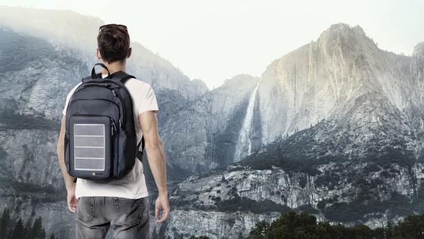 Ein Mann mit Rucksack blickt auf einen Wasserfall in einer bergigen Landschaft.