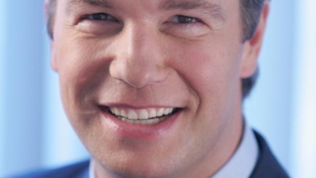 Ein lächelnder Mann im Anzug mit roter Krawatte vor blauem Hintergrund.