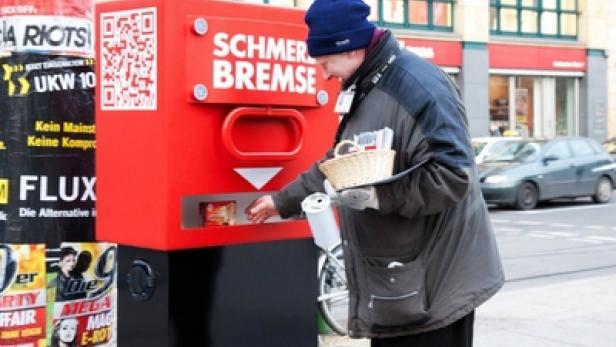 Ein Mann befüllt einen roten Automaten mit der Aufschrift „Schmer Bremse“ auf einer Straße.