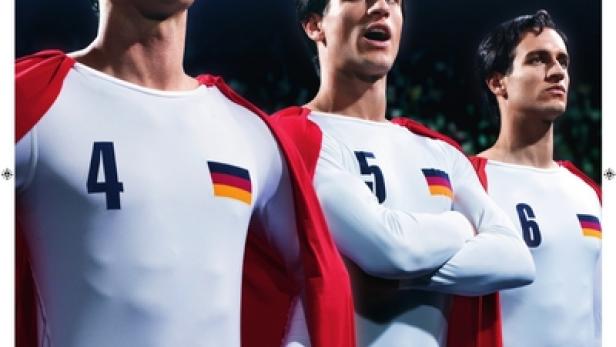 Drei Fußballspieler in Trikots mit Deutschlandflagge und roten Umhängen stehen auf einem Fußballfeld.