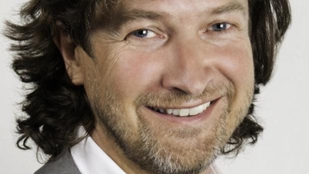 Ein lächelnder Mann mit braunen Haaren und Bart trägt ein graues Sakko.