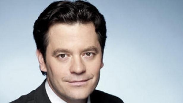 Ein Mann mit dunklen Haaren und Anzug vor einem hellblauen Hintergrund.