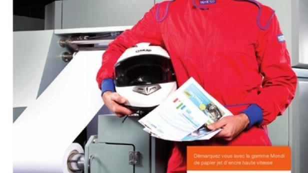 Ein Rennfahrer in rotem Overall hält einen Helm und bedrucktes Papier vor einer Druckmaschine.