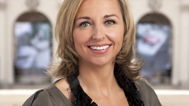 Porträt einer lächelnden Frau mit blonden Haaren vor einem Gebäude.