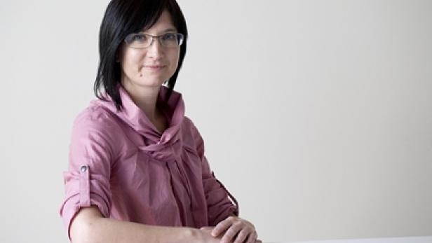 Eine Frau mit Brille und dunklen Haaren in einem rosafarbenen Hemd sitzt an einem Tisch.