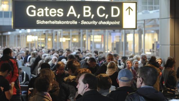 Eine Menschenmenge steht unter einem Schild mit der Aufschrift „Gates A, B, C, D“ und „Sicherheitskontrolle“.