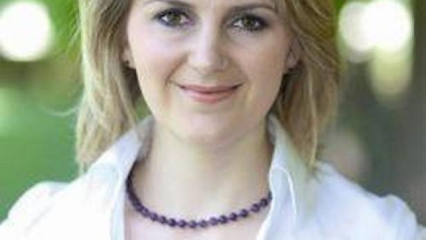 Eine Frau mit blonden Haaren und einer lila Halskette lächelt in die Kamera.