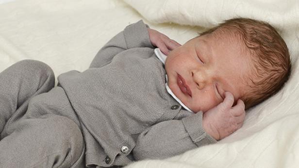Ein neugeborenes Baby schläft in einem grauen Strampler auf einer weißen Decke.