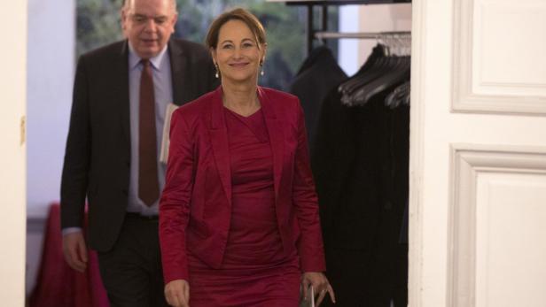 Eine Frau in einem roten Hosenanzug betritt einen Raum, gefolgt von einem Mann im Anzug.