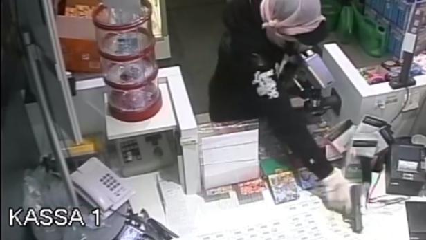 Eine maskierte Person mit einer Pistole steht an der Kasse 1.