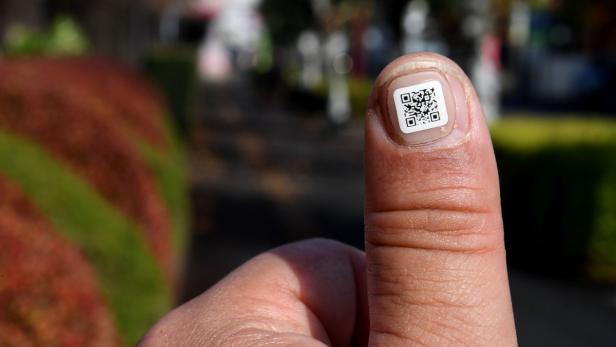 Auf dem Fingernagel einer Person befindet sich ein aufgeklebter QR-Code.