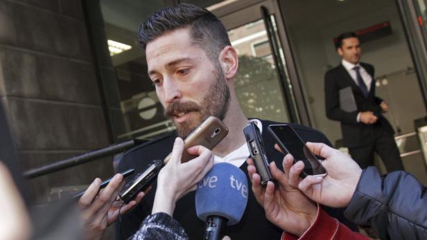 Ein Mann mit Bart wird von Reportern mit Mikrofonen und Handys umringt.