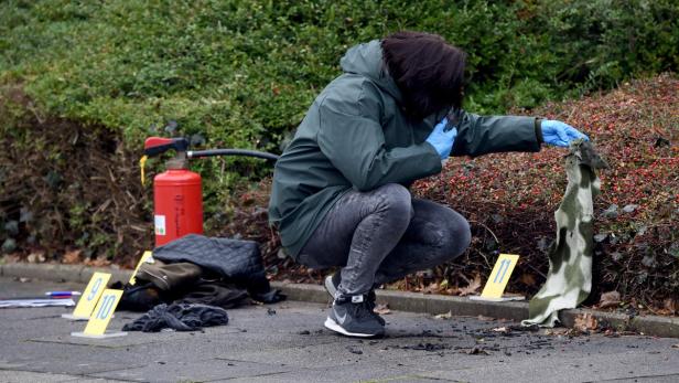 Eine Person in Schutzkleidung untersucht einen Brandort mit Beweismarkierungen und einem Feuerlöscher.