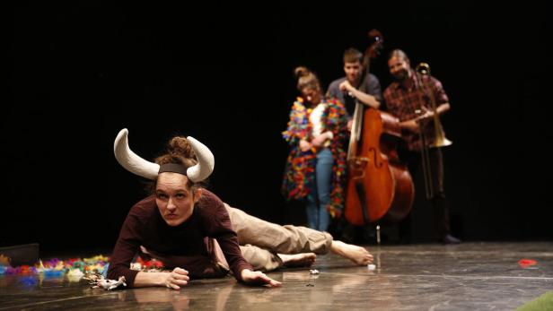 Eine Frau mit Hörnern liegt auf einer Bühne, im Hintergrund Musiker.