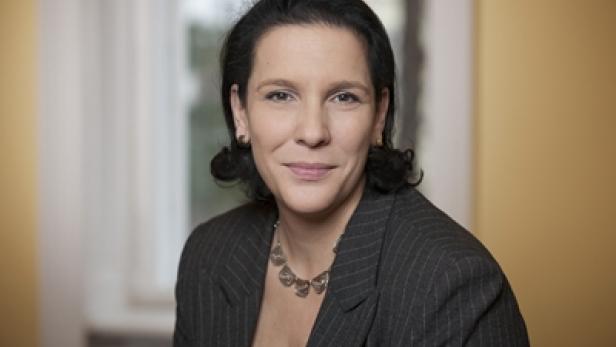 Eine Frau mit dunklen Haaren und einem grauen Blazer lächelt in die Kamera.
